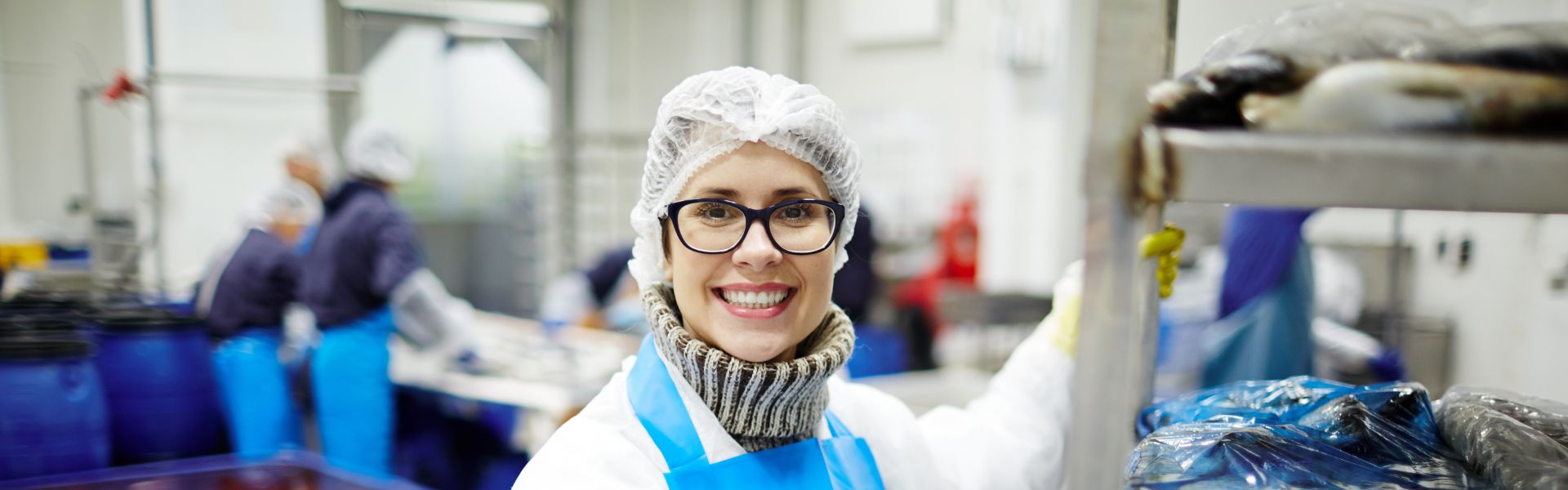 Worker with apron and hair net smiles as they pose for a picture beside a rack of fish.